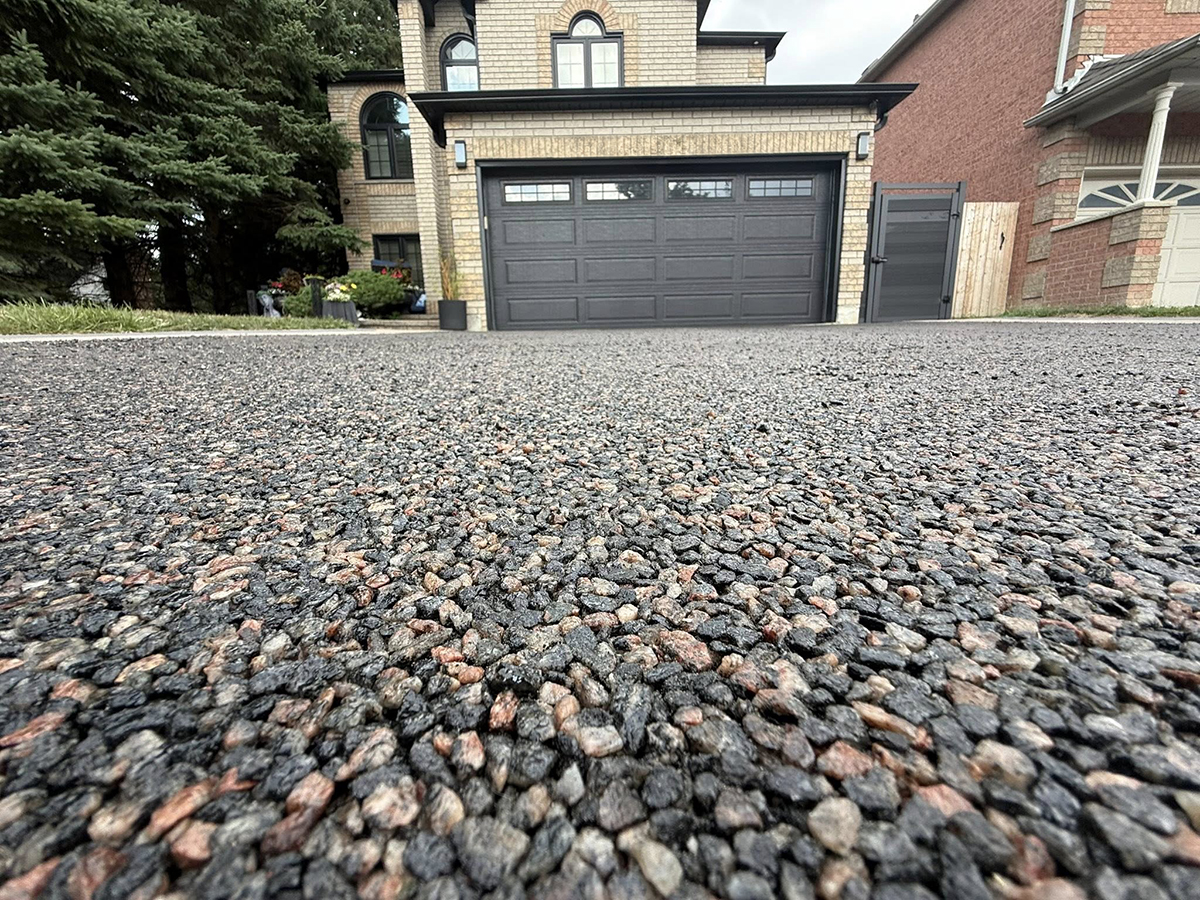 Close-up of rough asphalt driveway leading to a modern brick house with a closed dark garage door.