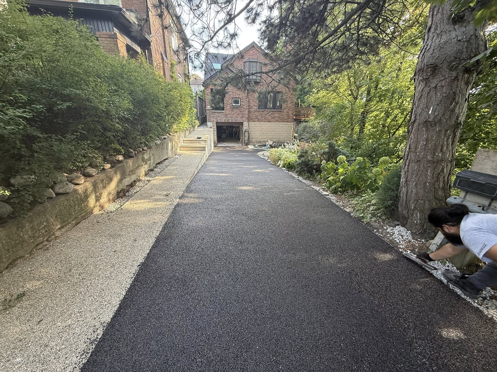 Man smoothing newly paved driveway beside a brick house surrounded by greenery and trees.