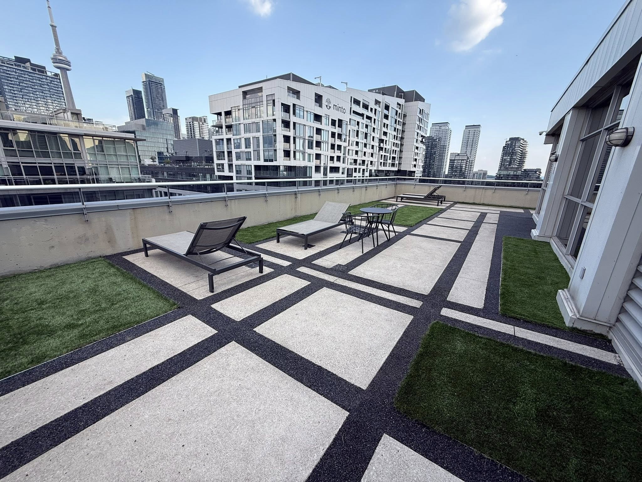 Rooftop terrace with lounge chairs, table, chairs, green grass patches, and city buildings including CN Tower.