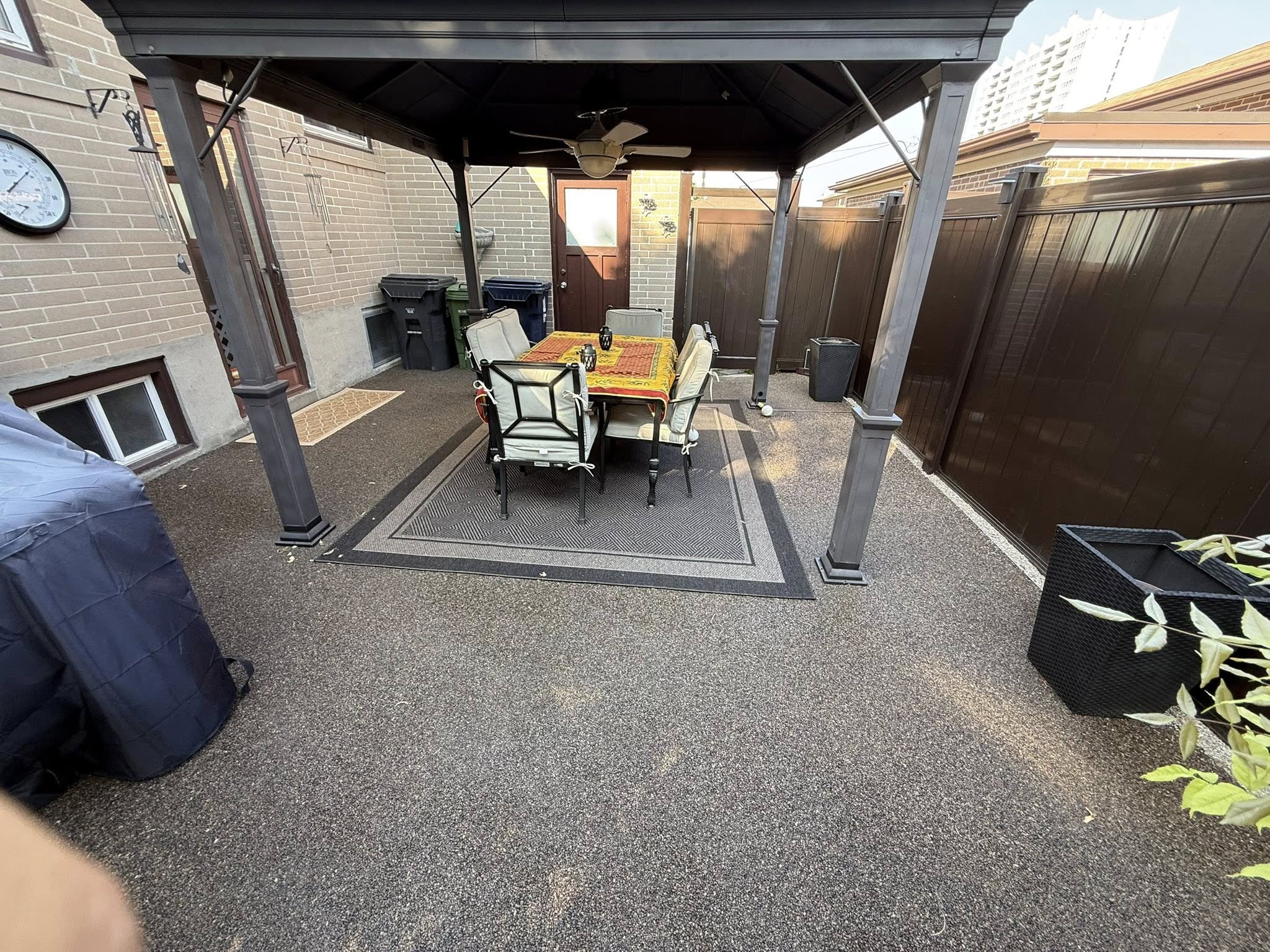 Covered patio with dining table, chairs, ceiling fan, and a brick wall with door and clock.