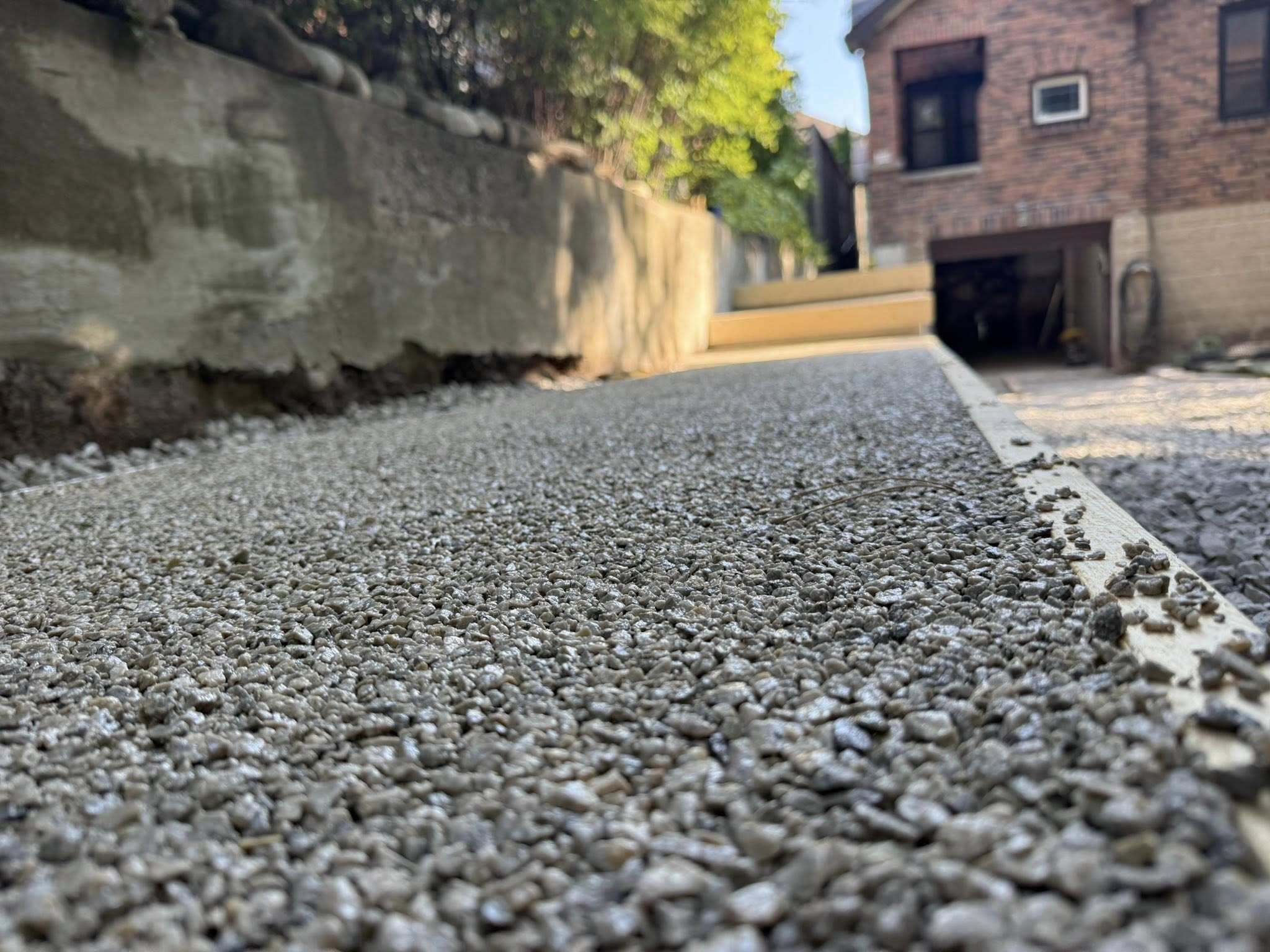 Close-up of wet concrete with aggregate set for a driveway near a brick house with garage.