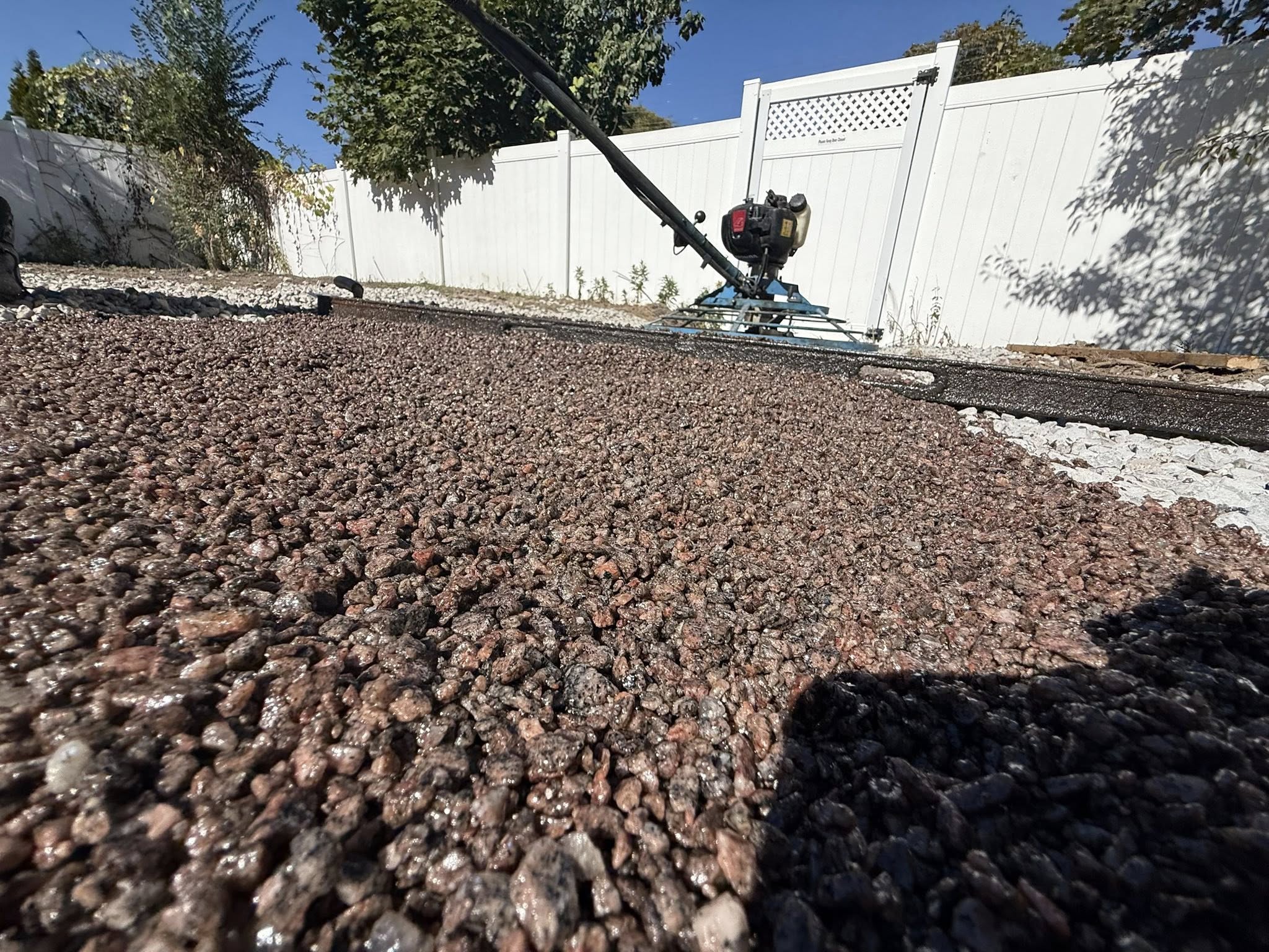 Close-up of reddish gravel surface with a blue concrete power trowel near a white fence and trees.